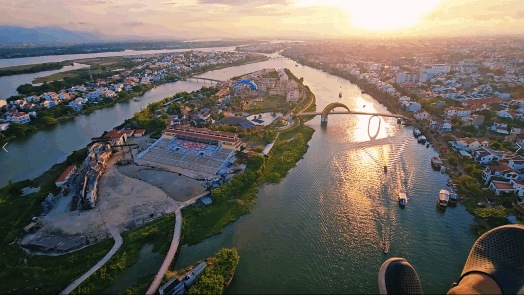 Aerial sunset view over the Thu Bon River and Hoi An Impression Theme Park, with a paraglider flying above the city. The glowing sun reflects on the water as boats glide below and a pedestrian bridge forms a circular “O” shape. A thrilling paragliding experience capturing Hoi An’s charm from above